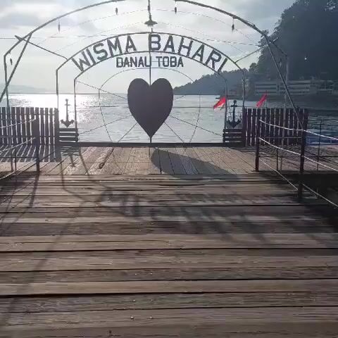 This is a video of an outdoor scene, likely a pier or boardwalk, with a body of water visible in the background. The foreground is dominated by weathered wooden planks, suggesting an aged structure. In the midground, there is an arched metal structure with the words "WISMA BAHARI" and "DANAU TOBA" inscribed on it, framing a large, dark heart shape. Flanking this archway are two dark wooden fences, each adorned with an anchor motif on its inner side. To the right, two small red flags are attached to a railing. The water in the background is calm and reflects the sunlight, indicating it is daytime. Hills or mountains covered in greenery can be seen on the far side of the water. The overall atmosphere appears serene and tranquil. There are no people visible in the video.