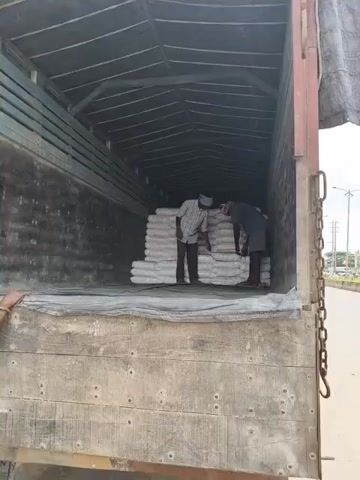 Two men are inside the back of a truck, which is loaded with sacks of white material. The truck is parked on the side of a road, and there is traffic visible in the background. The men appear to be unloading the sacks. The man on the left is wearing a plaid shirt and dark pants, and the man on the right is wearing a gray t-shirt and shorts. Both men are wearing turbans. The sacks are stacked neatly inside the truck, and they appear to be heavy. The truck is covered with a tarp, and the sides of the truck are made of wood. The road is paved, and there are buildings and trees in the background.