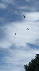 Five military helicopters fly in V-formation through partly cloudy skies.