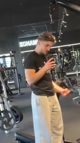 A young man is standing in a gym, facing a mirror. He is wearing a black t-shirt and light gray sweatpants. He has brown hair and a beard. The man is flexing his right bicep while holding a smartphone in his left hand, apparently taking a selfie or recording a video. The background shows various gym equipment, including weight machines and benches, and a sign that reads "Performance". The overall lighting in the gym appears bright.
