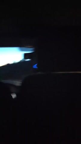 The video is shot from the back seat of a moving car at dusk. The driver is visible as a dark silhouette. The dashboard is dimly lit with red lights. Through the windshield, one can see a glimpse of the sky transitioning from blue to a darker hue, with trees visible on the horizon. The road ahead is illuminated by the car's headlights. A blue ring-shaped object is hanging from the rearview mirror.