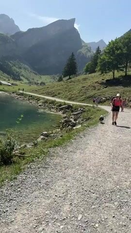 The video begins with a shot of a gravel path next to a clear, green lake. Mountains rise in the background, covered in trees and rocky cliffs. A few people and a dog are walking on the path. The camera pans to the left, revealing more of the lake and the surrounding landscape, including some small buildings on the shore. The water is transparent, allowing the rocky bottom to be seen. The trees are lush and green, and the sky is a clear blue. The video continues to show the lake and mountains from various angles, emphasizing the natural beauty of the location. The overall impression is one of a serene and picturesque mountain lake scene.