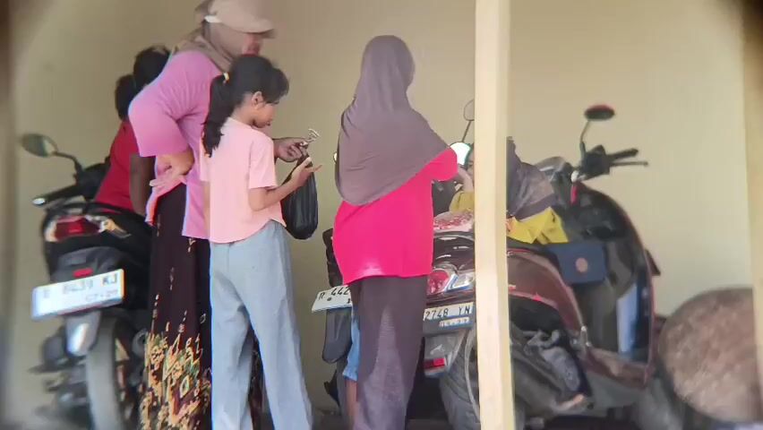 Four people, three adult females and one young female child, are standing under a covered outdoor area. Two motorcycles are parked behind them. The adult females are wearing headscarves, and one is wearing a pink hijab, another a grey hijab, and the third a lighter colored hijab and a sun hat. The young female child is wearing a pink shirt. The people appear to be interacting with each other and looking at a cell phone. One of the adult females is laughing. The environment appears to be a hot, sunny day.