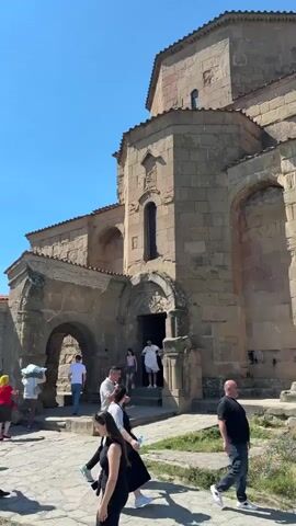 The video shows the exterior of an ancient stone church with a large arched entrance. Several people are gathered around the entrance and on the paved area in front of the church, some appearing to be tourists. A large tree with green foliage provides shade to a group of people on the left side of the frame. The background reveals a mountainous landscape under a clear blue sky with scattered clouds. The ground is paved with irregular stones.