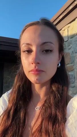 A young woman with long, dark, wavy hair is shown in a close-up shot. She is wearing a white top with puffed sleeves and a silver necklace with a CD pendant. She has on hoop earrings and appears to be outdoors, with a stone wall and wooden siding in the background. The lighting suggests it is either sunrise or sunset, casting a warm glow on her face. She makes several subtle facial expressions, including looking down, closing her eyes briefly, and looking directly at the camera with a slight smile.