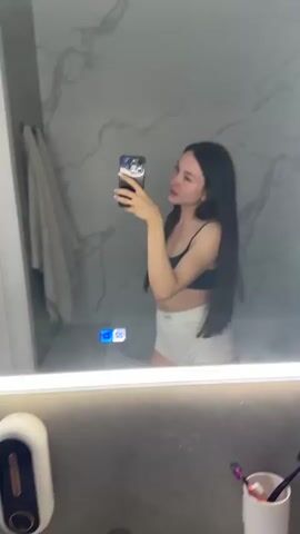 A young woman with long dark hair is taking a selfie in a bathroom mirror. She is wearing a black sports bra and white shorts. She is leaning on her hands, which are on the edge of the sink. The mirror is illuminated by a bright light around its edges. The bathroom walls are covered in marble-patterned tiles. There is a white towel hanging on a rack to the left of the mirror. Below the mirror, there is a white sink with a silver faucet. To the right of the sink, there is a white cup holding two toothbrushes.