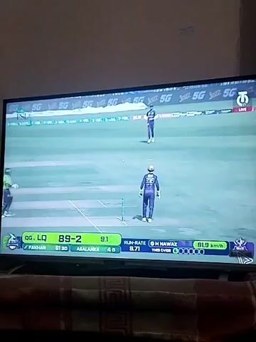 A cricket match is being shown on a television screen, likely indoors, with the TV resting on dark-colored furniture. The screen displays a green cricket field with three players visible: one bowling, one batting, and one standing near what appears to be the umpire's position.

On-screen graphics provide match information:
- "LIVE" is displayed in a red box at the top right.
- The teams "QG, LQ" are shown with the score "89-2" after "9.1" overs.
- Player names and their individual scores are "FAKHAR" and "ASALANKA".
- The "RUN-RATE" is "9.71".
- The bowler's name "M NAWAZ" is visible, along with his bowling figures "0-18 1.1".
- The progress of "THIS OVER" is represented by "000000".
- A bowling speed of "91.9 KM/H" is also displayed.
- A "PSL" (Pakistan Super League) logo is visible on the top left, and multiple "5G" logos are present in the background of the cricket field.