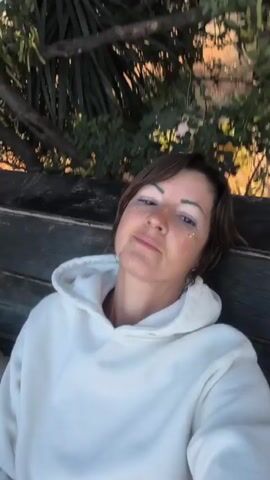 A woman with short brown hair, wearing a white hooded sweatshirt, sits on a wooden bench outdoors in a selfie video. She has subtle glitter on her upper cheekbones and a nose ring. She looks directly at the camera, blinking and offering slight smiles, occasionally shifting her head. Out-of-focus trees and foliage are visible in the background.