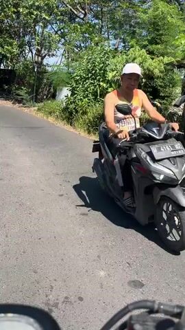 A man wearing a white cap and a sleeveless shirt is sitting on a black motorcycle. The motorcycle is parked on an asphalt road. The man is looking to his right and appears to be talking. He is holding the handlebars of the motorcycle. Behind the man, there are green trees and foliage. The lighting suggests it is daytime and sunny. The road appears to be a rural or suburban area.