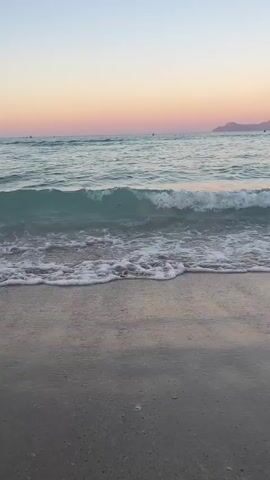 The video shows a close-up view of a calm beach at sunset. Gentle waves roll onto the sandy shore. The sky is a gradient of soft pink, orange, and blue. In the distance, a faint silhouette of land can be seen. There are a few indistinct figures in the water far offshore.