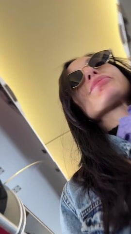 A woman wearing sunglasses is on an airplane. She is holding a boarding pass and a passport and smiling for the camera. The plane cabin is visible in the background.