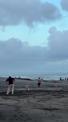 The video shows a beach scene on a cloudy day. There are several people on the dark sand. A woman in a black shirt and light-colored pants is standing with her hands on her hips, then raises her arms. Near her, two children, both wearing pink, are walking on the beach, and a man in a pink floral shirt is kicking a ball. Other people can be seen further down the beach, some walking along the shore. The ocean is visible in the background, with gentle waves. The sky is overcast with large, gray and white clouds.