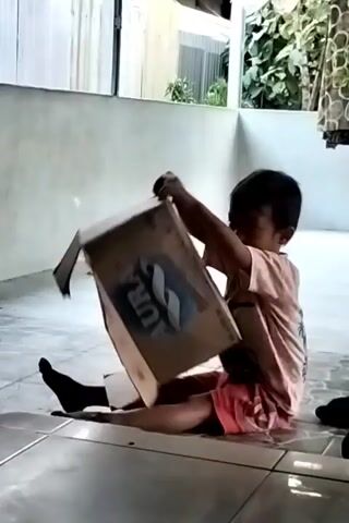 A young child sits on a tiled floor, playing with a cardboard box. The child is wearing a pink t-shirt and red shorts. The box has a logo that reads "AURA". The child is holding a stick or a tool and appears to be trying to poke or open the box. The background is out of focus but shows a wall and some greenery.