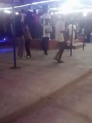 The video shows a group of people at an outdoor party under a tent-like structure.  A crowd of people are gathered in the background, some standing on a low platform.  In the foreground, several people are moving around, and one woman in an orange shirt and black pants walks across the frame.  The lighting includes bright overhead lights, and some colored lights that cast a purple and green glow. The ground appears to be concrete.  Music is playing.