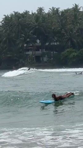 A woman stands up on a surfboard and rides a wave toward the beach. She has her arms out to the sides and is looking forward. Behind her, several people on surfboards are paddling in the water. Palm trees and buildings are visible in the background. The sky is overcast.