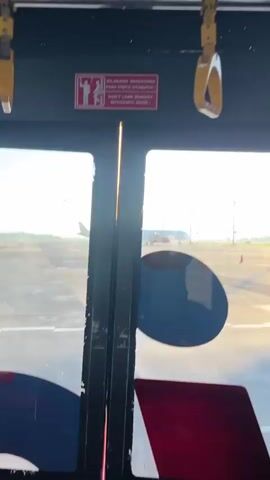 The view from inside a bus, with an open door, shows a window with a view of a plane on a runway. The bus is black with a yellow plastic hand grip hanging from the ceiling. A sign in Indonesian and English warns passengers not to lean against the automatic door. Outside the bus, the sun is shining brightly on the tarmac. The tail of a white airplane is visible, and as the bus moves, more of the plane, with the registration PK-GAV, comes into view. A person in a blue shirt and watch is holding the metal pole on the bus door.