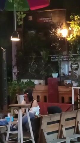 The video opens with a woman sitting at a small wooden table outdoors at night. She is wearing glasses, a black top, and jeans. She is holding a phone or small object in her hands and appears to be looking down at it. Behind her, there is a wall with some plants and string lights. There is also a sign with text on it, and an umbrella is visible hanging above. To her right, there are wooden benches and more plants. The overall atmosphere appears to be a casual outdoor dining or lounge area.