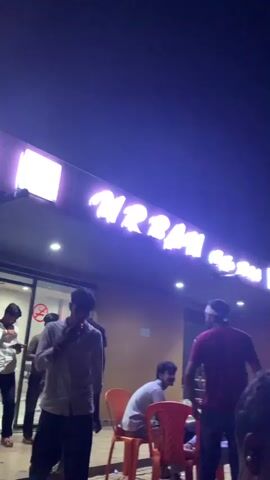 The video captures an outdoor scene at night, likely a restaurant or roadside eatery, illuminated by a neon sign and ambient lighting. Several people are visible, some standing and others seated at tables. The neon sign displays text in an unidentifiable script, glowing in shades of purple and white. Orange plastic chairs are arranged around tables, and palm trees are dotted around the area, suggesting a tropical or subtropical location. The overall atmosphere appears casual and social, with people engaged in conversation or simply present in the environment. An Indian flag is visible on a flagpole in the background. The video is shot from a slightly low angle, focusing on the interactions and the environment.