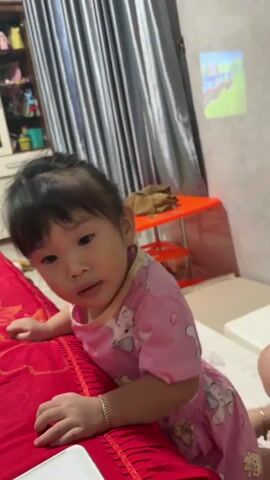 The video begins with a close-up of a young child wearing a pink shirt with cartoon characters. The child is looking at the camera with a neutral expression. The background is blurry but appears to be an indoor setting with curtains and furniture. The camera then moves closer to the child, and their face fills the frame. The child blinks and their mouth is slightly open. The video continues with a series of quick cuts, with the child's face appearing in and out of focus. There are brief glimpses of a person's hand and another person in the background. The environment remains consistent, with the child in the foreground and blurry objects and furnishings in the background. The video ends with the child's face blurry and out of focus.