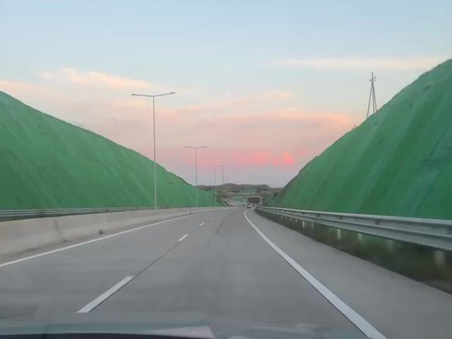 A daytime, front-facing, outdoor shot from a car shows a multi-lane highway with green netting-covered hills on either side. A white guard rail separates the two sides of the highway. Above, the sky is light blue with a band of pink clouds in the distance. The road ahead curves to the right, and the sun is visible in the distance.
