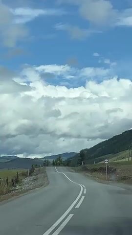 The video shows a road ascending a hill in a rural landscape. The sky is blue with large white clouds. There are mountains in the distance. The road has a white line dividing the lanes, and white dashed lines in the lane. There is a roadside barrier on the left side of the road, and some trees on the right side. There is also a traffic sign on the right side of the road.