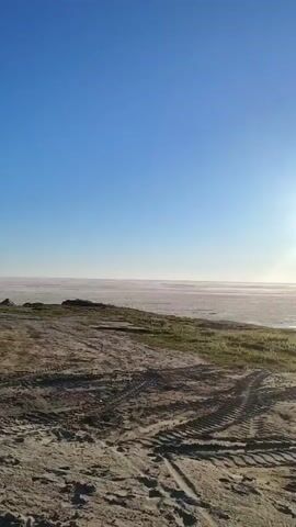 The video is an outdoor, vertical, landscape shot. The sky is a clear blue with the sun shining brightly in the upper right portion of the frame. The sun creates a lens flare effect. In the foreground, there is rough, sandy terrain with tire tracks visible. A patch of green grass is present to the left and right of the sandy area. The background features a flat, hazy horizon line that appears to be a body of water or a very flat, expansive landscape. The overall impression is of an open, natural environment under a bright sky.