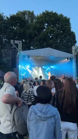 A concert is underway outdoors. A band is performing on a stage that is illuminated by bright lights, primarily blue and purple. The stage is set up under a white canopy and is surrounded by metal scaffolding. A crowd of people, mostly young adults, are gathered in front of the stage, watching the performance. The lighting casts a hazy glow, and some of the crowd are facing the stage. The time stamp indicates the video is just starting.