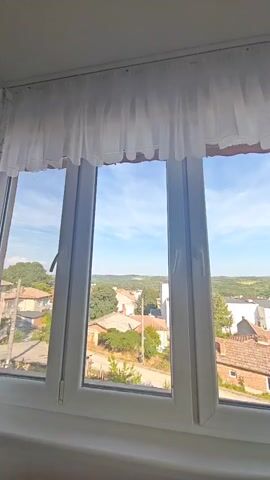 The video opens with an indoor shot through a white-framed window, revealing a sunny village scene with houses and distant green hills under a bright sky. The camera then pans upwards and outwards, moving beyond the window frame to show an expansive view of a bright blue sky with faint clouds, green rolling hills, and the distant tops of houses and trees. The entire scene is bathed in clear daylight.
