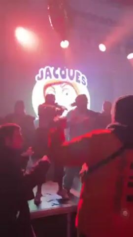 The video shows a crowded live music performance from the perspective of someone in the audience. A stage is visible with the word “JACQUES” displayed on a large screen behind the performers. The stage is lit with red and blue lights.  Several individuals can be seen on the stage, seemingly performing, but their actions are partially obscured by the audience. People in the audience are moving, dancing, and raising their hands, indicating they are enjoying the performance. A red heart-shaped balloon floats near the top of the frame. The word “HIPPODROME” is visible above the stage.