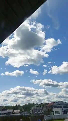 The video shows a time-lapse of clouds moving across a blue sky. In the foreground, there is a partial view of a dark overhang, likely part of a roof or structure. Below the sky, a residential area with houses and trees is visible. The houses have various roof styles and colors, suggesting a suburban or rural setting. The clouds are white and fluffy, and their movement indicates a moderate wind. The overall scene is bright and sunny, characteristic of daytime. There are no people or explicit actions captured in the video, only the natural movement of the clouds and the static view of the landscape.