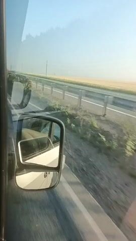 The video shows a view from inside a vehicle, looking out the side window. A side mirror is visible, reflecting part of the vehicle's interior and exterior. Outside the window, a highway stretches into the distance, bordered by a guardrail and a field of crops. Trees are visible in the background, and the sky is clear and blue. The video appears to have been filmed while the vehicle was in motion, as indicated by the blur of the passing scenery. There are no people clearly visible in the video.