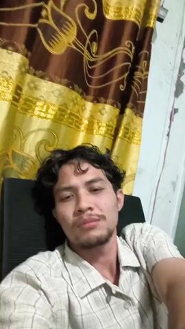 This is a selfie-style video clip featuring a young man with curly dark hair and a light beard. He is wearing a light-colored, plaid button-down shirt. He is seated in what appears to be an office chair with a dark back. Behind him, there is a curtain with a brown and gold patterned design. The lighting suggests it is daytime, though the exact time is not specified. The man makes a peace sign with his right hand and waves it slightly at the camera. His expression is neutral to slightly pensive. The scene appears to be indoors, likely in a home or office setting in Pulodang, Indonesia. There is no visible text in the video.