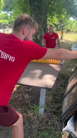 Two men are playing table tennis outdoors. The man in the foreground, wearing a red shirt and burgundy shorts, is facing away from the camera. The man in the background, also in a red shirt and grey shorts, is facing the camera and appears to be returning a serve. The table tennis table is set up on a grassy area with trees in the background.