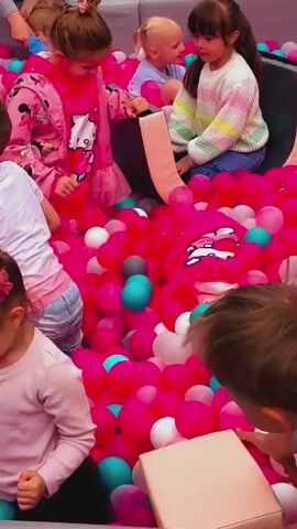 The video shows multiple young children playing in a large, gray, soft-sided ball pit filled with plastic balls in various shades of pink, fuchsia, white, light blue, and turquoise. A mint green, curved soft play structure is positioned in the center of the pit. Several children are actively engaging with the balls, sitting, crawling, and digging through them. One girl is wearing a pink hoodie with a Hello Kitty design, another a pink jacket with Mickey Mouse characters, and a third in the foreground wears a plain light pink long-sleeve shirt. A boy in a white t-shirt and jeans is seen crawling near the mint green structure. An adult's hands are visible at various points, seemingly supervising or assisting the children. The children appear happy and engaged in the activity.