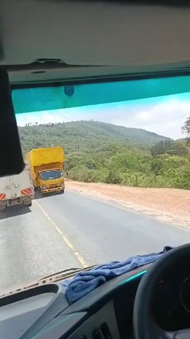 The camera is inside a vehicle, likely a truck or bus, driving along a paved road through a hilly, green landscape. A yellow truck covered in a yellow tarp passes in the opposite direction. A red and white van also passes. Further ahead, another truck can be seen. The sky is partly cloudy. A blue cloth sits on the dashboard. The speaker seems to be discussing the other vehicles passing them, mentioning technology.