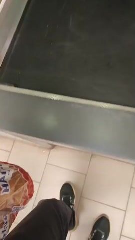 The video shows a close-up shot of a supermarket checkout counter. A person wearing a green vest is standing behind the counter, and their hands are visible. In front of them, on the black conveyor belt, are two items: a packaged food item with Russian text that appears to be noodles or vermicelli, and a white plastic tray holding it. The hands of another person, presumably the customer, are holding a plastic bag of bread. The customer is placing the bread and the noodle package on the conveyor belt. The background shows shelves stocked with various products and a part of a cash register. The lighting is typical of a commercial setting.
