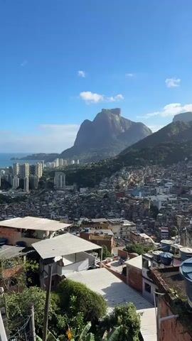 A high-angle, vertical shot captures a bustling city nestled amongst verdant mountains under a bright blue sky with scattered clouds. The foreground reveals a densely packed residential area with numerous rooftops and a blue water tank labeled "FORTLEV" on the right. In the midground, the city stretches out, filled with countless buildings and structures, some appearing as favelas climbing the hillsides. The background is dominated by majestic mountains, with a prominent, flat-topped peak visible on the left and other rugged peaks extending to the right. A glimpse of the ocean can be seen in the far left background, beyond a cluster of modern high-rise buildings. The camera slowly pans to the right, revealing more of the sprawling cityscape and the dramatic mountain landscape.