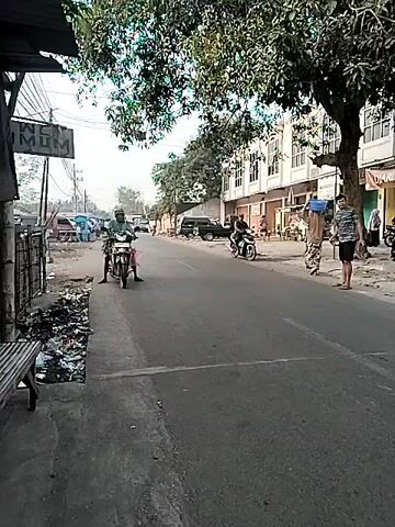 A street scene unfolds, featuring several people and vehicles. On the left side of the road, a person on a motorcycle is stationary, facing the camera. To the left of the road, a bench is visible next to a pile of trash. Further down the road, on the right side, two individuals are standing and another person is on a motorcycle. Across the street, several buildings line the road, with some appearing to be shops with closed shutters. A large tree with dense foliage extends into the frame from the top right, providing shade over a portion of the street. The road itself appears to be paved and has white lines marking a pedestrian crossing. The overall environment suggests a public street, possibly in a town or city, with moderate activity.