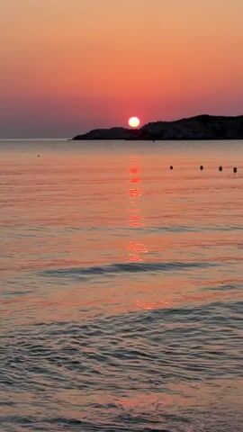 The video shows a serene beach scene at sunset. The sun is a glowing orb setting over a distant island, casting a vibrant orange and pink hue across the sky and reflecting a shimmering path on the calm ocean water. Gentle waves lap at the sandy shore in the foreground. A few buoys are visible floating on the water. The overall atmosphere is peaceful and tranquil.