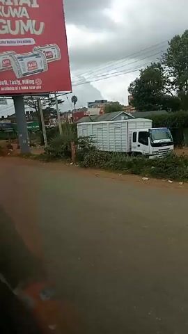 A view of a street in Kenya with a large billboard advertising bread on the left. Cars and a bicycle pass on the street. There is a white truck parked in the background. A person is walking along the side of the road. The sky is overcast.