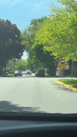 The video shows a view from inside a car looking out at a residential street with trees lining both sides. The street has a stop sign at the intersection with the words "CROSS TRAFFIC DOES NOT STOP" below it. Several cars are visible in the distance approaching the intersection. The sky is blue with some light clouds. The video is shot during daylight hours. The cars are moving slowly.