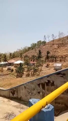 The video shows a view of a man-made reservoir or dam. The camera is positioned on what appears to be a walkway or bridge overlooking the water. A yellow and blue railing runs along the side of the walkway. The water is calm and a light brownish-green color. The surrounding landscape is hilly and covered with trees, some of which appear to have been cleared on the slopes. In the distance, there are a few buildings visible. The sky is clear and blue with a few scattered clouds. The video appears to be taken during daylight hours. There are no people visible in the video.