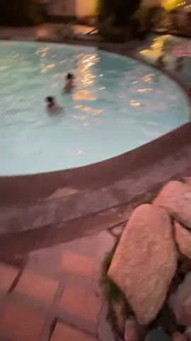 The video opens to a wide shot of a swimming pool at dusk. The sky is a vibrant gradient of pink, orange, and purple. Several people are in the pool, including children and adults. In the background, palm trees and a building with lights are visible. The camera pans to the right, showing more of the pool area and the sunset. The focus then shifts to two palm trees with the sunset behind them. The last shot shows a person standing by the pool with the colorful sky above.