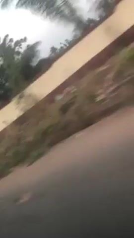 The camera is moving rapidly along an asphalt road, filming from a low angle. On the right, there's a low, tan-colored wall bordering grassy ground. Above the wall, there are trees and an overcast sky. The video is shaky due to the speed and movement of the camera.