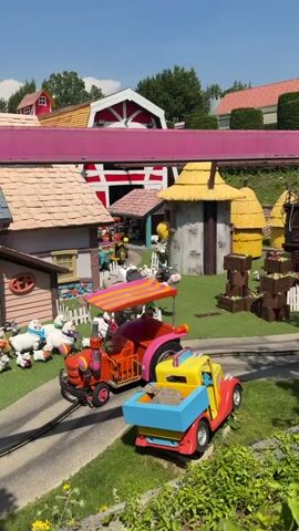A bright, sunny day at an amusement park. A small, red and orange tractor, possibly a ride for children, is on a track. It has a striped canopy and a driver. A yellow and blue toy truck with a load of dirt is behind it on the same track. In the background are cartoonish buildings with colorful roofs and walls, made to look like farm structures. There are also stuffed animals resembling farm animals like sheep and a cow scattered around the scene. The overall atmosphere is playful and whimsical, suggesting it's a themed area for young children.