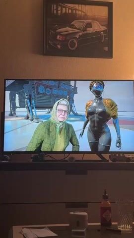 A television screen displays a scene from a video game, featuring an older woman in a green jacket and headscarf on the left, and a futuristic female robot in a metallic suit with a golden jacket on the right. Above the TV, a framed picture depicts a dark Mercedes-Benz W124 car in an urban setting. Below the TV, on a dresser or media console, there is a red bottle of Tabasco sauce, a clear glass with liquid, and a white cylindrical object. A lamp on the right casts a warm glow, suggesting it is evening or night. The wall behind the TV has light-colored, textured wallpaper.