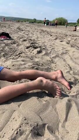 The video shows a child lying on a sandy beach. The child is wearing red, white, and blue swim trunks. The child is face down in the sand with their arms covering their face. There are some shoes and a black bag near the child. In the background, other people are visible on the beach and in the water. The sky is blue with some clouds.