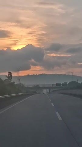 This is a vertical, eye-level shot looking down an empty, gray asphalt road towards a distant bridge overpass. The sun is setting or rising behind a line of hazy, dark green hills in the background, casting a warm orange and yellow glow through patchy, gray clouds. On the left side of the road, a shallow grassy embankment is visible, with a tall metal power line tower. On the right side of the road, there are more power line towers and the road curves slightly to the right. The road is divided by a solid white line on the left and a broken white line on the right. There are no people visible in the shot, and the only visible objects are the road, the power line towers, the bridge, and the hills.