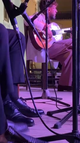 The video captures a low-angle view of a musical performance. In the foreground, the legs and feet of two musicians are visible, clad in dark trousers and dress shoes. One musician wears black dress shoes with laces, while the other has woven black shoes. Microphones on stands are prominently positioned in the shot. In the background, a guitarist with curly hair is seated on a chair, playing an acoustic guitar. He is wearing a white shirt and light-colored trousers. The stage or performance area is lit with purple and pink lighting, casting a vibrant glow on the scene. The background environment appears to be outdoors, with a building structure visible behind the guitarist. The overall scene suggests a live musical event.
