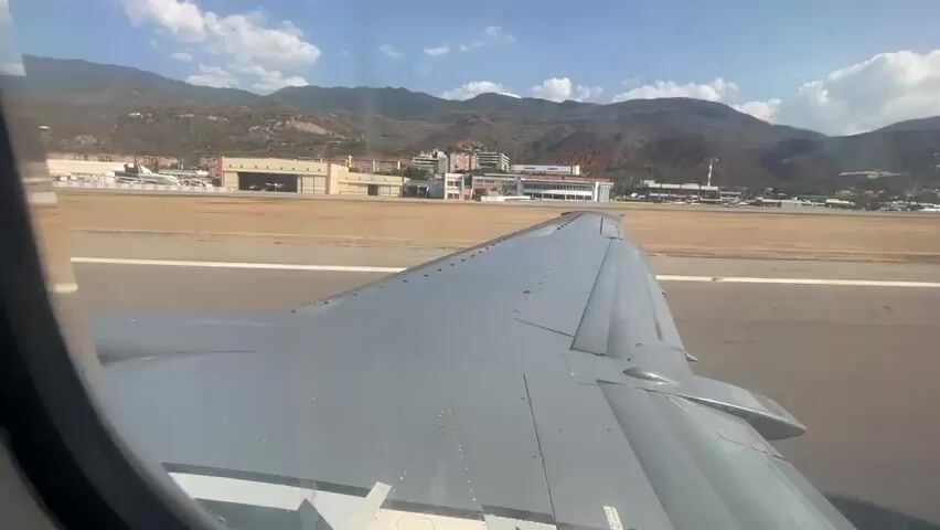 From the perspective of an airplane window, an aircraft accelerates on a runway, its wing prominently visible. The runway extends towards a backdrop of buildings and mountainous terrain under a bright, partly cloudy sky. Several other airplanes are visible parked near airport infrastructure.